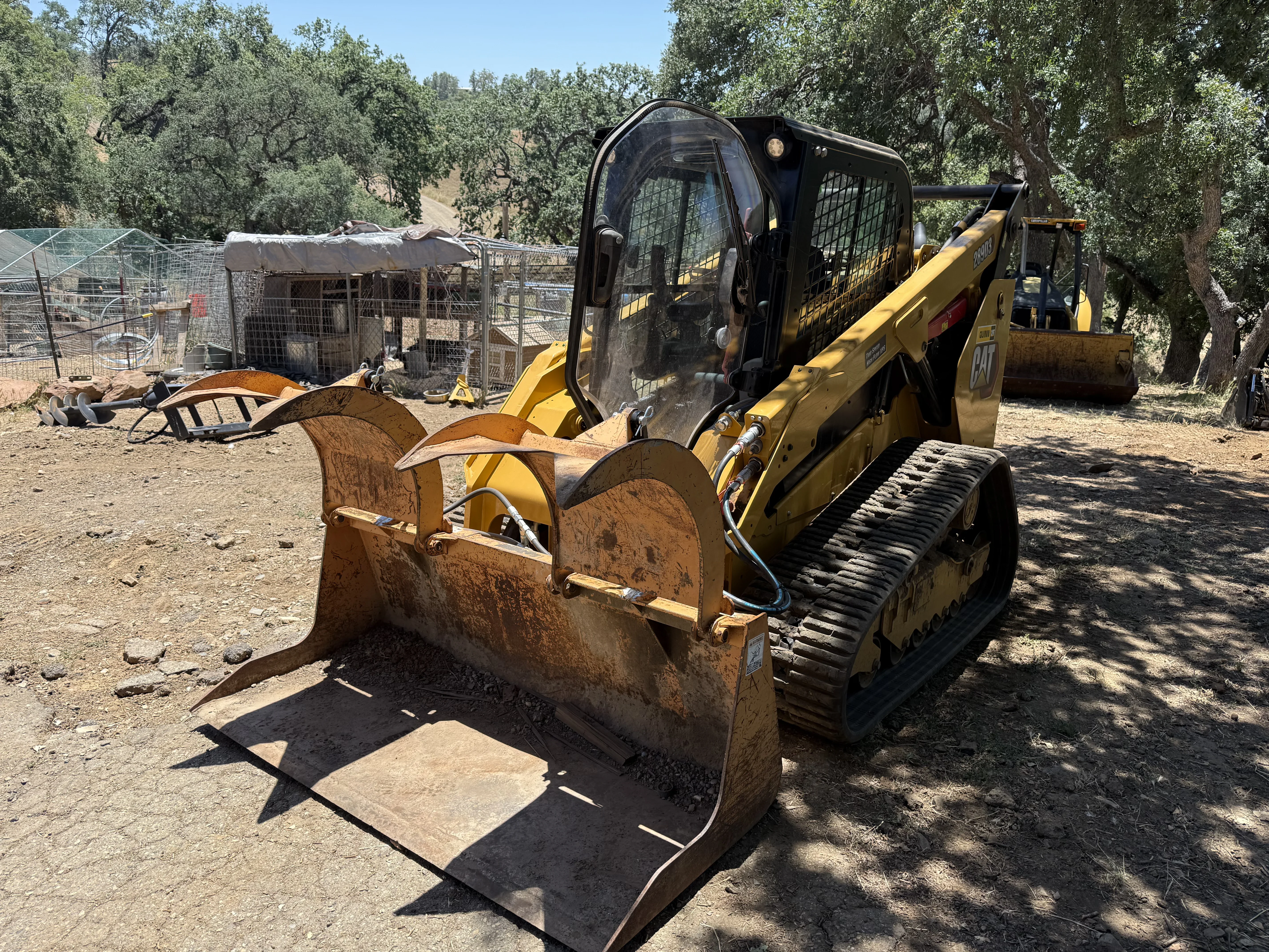 Skid steer loader with grapple attachment clearing debris on rural property with oak trees