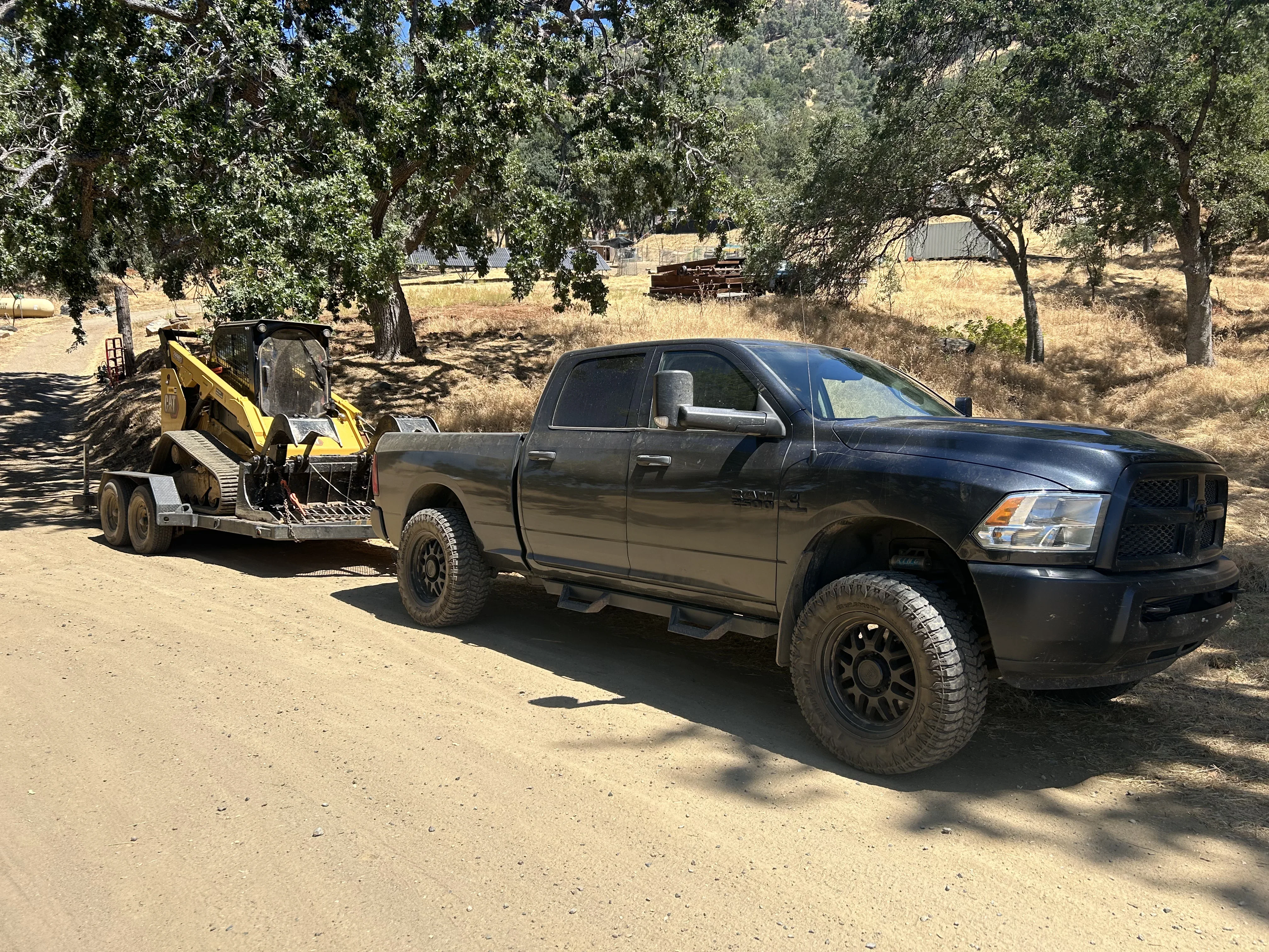 Skid steer loader with debris bucket parked beside pickup truck on cleared dirt lot with oak trees